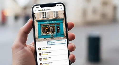 smartphone-affiche-fiche-google-business-profile Un smartphone tenu en main affiche une fiche Google Business Profile complète d’un commerce local à Arras, avec des avis positifs visibles, une photo de vitrine et un bouton « Itinéraire » en surbrillance.